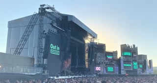 Die Q Stage unter freiem Himmel mit Dach, Lautsprechern und Videowänden mit der Aufschrift „Out in the Green“, davor Publikum.
