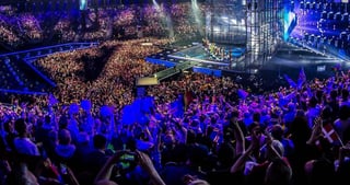 Publikum mit Fahnen in einer vollen Arena beim Eurovision Song Contest, im Hintergrund die beleuchtete Bühne.